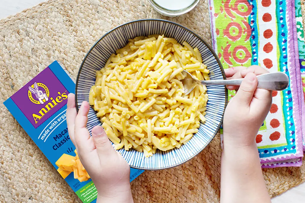 Hands holding and stirring a bowl of mac and cheese next to a box of Annie's Macaroni And Classic Cheddar.