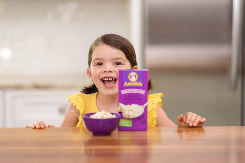 A young girl smiling sitting at a table with a box of Annies Mac & Cheese next to a bowl.